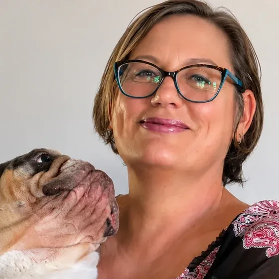 Rose, in glasses, smiling as an older bulldog in her arms stretches up toward her face